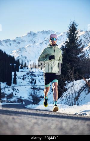 Mann Athlet, der auf der Straße im schneebedeckten Bergtal läuft, am sonnigen Wintertag. Joggen in Sportbekleidung in der Natur. Stockfoto