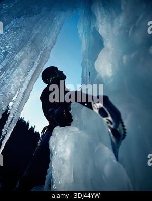Ein Mann in gelber Jacke klettert mit Eisaxt und Steigeisen an einer gefrorenen Eiswand am großen Wasserfall gegen blaue Ski in den Winterbergen. Sport Bergsteiger Stockfoto