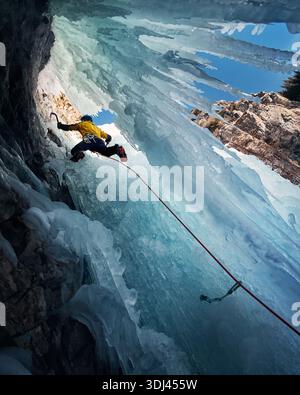 Ein Mann in gelber Jacke klettert an der gefrorenen Eiswand am großen Wasserfall r mit Eisaxt und Steigeisen gegen blaue Ski in den Winterbergen. Sport Mountain Stockfoto