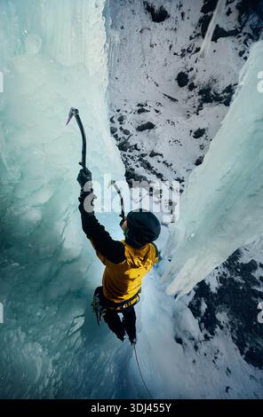 Ein Mann in gelber Jacke klettert an der gefrorenen Eiswand am großen Wasserfall r mit Eisaxt und Steigeisen gegen blaue Ski in den Winterbergen. Sport Mountain Stockfoto