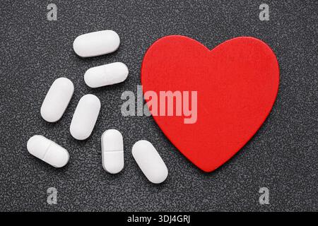 Weiße Pillen neben roter Herzform auf dunkler Oberfläche. Medizin und Gesundheitskonzept. Behandlungsunterstützung und Herzgesundheitsbewusstsein. Stockfoto