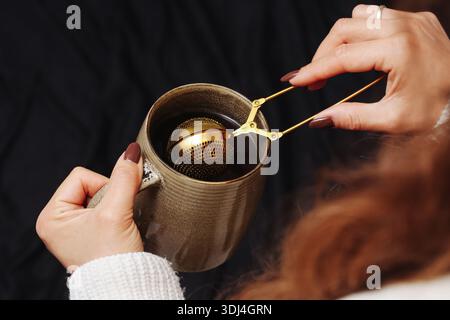 Zubereitung von grünem Tee mit goldenem Infuser. Weibliche Hände rührend Kräutergetränk in Metallbecher. Nahaufnahme des Heißtee-Zubereitungsprozesses. Stockfoto