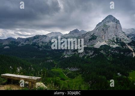 Eindrucksvolle, spitze Gipfel im Triglav Nationalpark, an einem stimmungsvollen, bewölkten Tag mit viel Licht und Fokus auf der vorderen Holzbank mit Aussicht Stockfoto