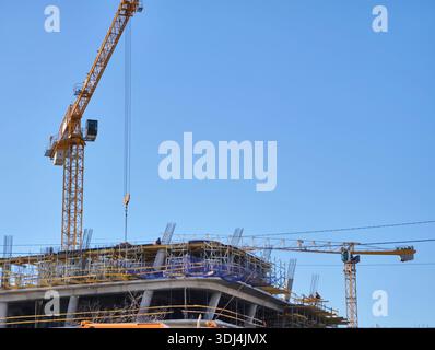Baukräne ragen über einem belebten Gelände unter einem klaren blauen Himmel hervor, was Fortschritte bei der Stadtentwicklung und architektonischen Innovation hervorhebt Stockfoto