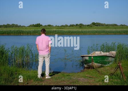 Rückansicht eines Mannes, der auf einem Flussufer neben einem alten Boot steht, das in einem dichten Schilfbett verankert ist. Stockfoto