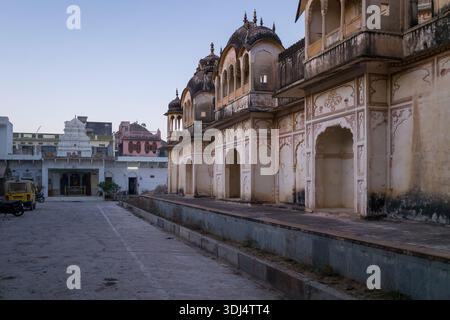 Pushkar, Indien - 18. März 2022: Der Innenhof des Sri Rangnath Swamy Temple Complex ist mit verwitterten Tempelgebäuden mit kunstvollen Bögen und Kuppeldächern, einem weißen Schrein, einem gelben Fahrzeug und einem Motorrad in der Abenddämmerung zu sehen. Stockfoto