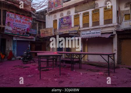 Pushkar, Indien - 18. März 2022: Mehrere geschlossene Geschäfte mit sichtbaren Schildern und Bannern sind entlang einer Straße mit rosa Holi-Pulver bedeckt, mit leeren Tischen im Vordergrund und einem Mann, der auf einem Motorrad sitzt. Stockfoto