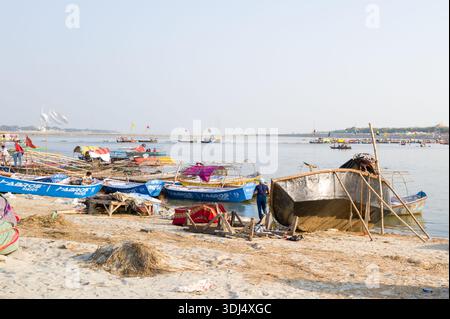 Prayagraj, Indien - 21. März 2024: Mehrere Menschen arbeiten an und um Boote herum, darunter ein großes umgedrehtes Boot, das repariert wird, entlang des Sandufers bei Sangam mit zahlreichen überdachten und nicht überdachten Booten, die in der Nähe verankert sind, und weitere Boote, die unter klarem Himmel auf dem Wasser zu sehen sind. Stockfoto