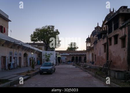 Pushkar, Indien - 18. März 2022: In einem Innenhof des Swamy Temple von Sri Rangnath werden ein geparktes Auto, ein großer Baum und Menschen gezeigt, die in der Nähe historischer Tempelgebäude mit Kuppeldächern und bogenförmigen Eingängen in der Abenddämmerung spazieren. Stockfoto