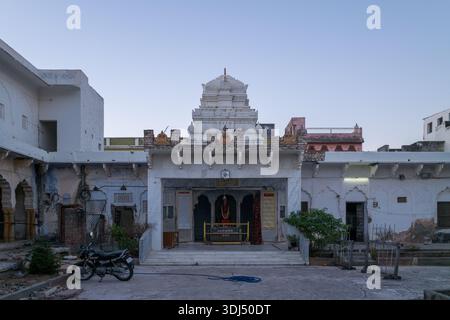 Pushkar, Indien - 18. März 2022: Der Innenhof des Swamy Tempels von Sri Rangnath wird mit einem zentralen weißen Schrein, kunstvollen architektonischen Details, einem geparkten Motorrad, kleinen Bäumen und umliegenden verwitterten Gebäuden in der Abenddämmerung gezeigt. Stockfoto