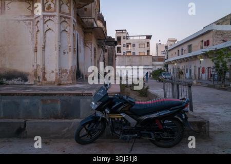 Pushkar, Indien – 18. März 2022: Neben einem verwitterten Tempelgebäude mit kunstvollen Bögen steht ein Motorrad, während Menschen und Fahrzeuge in der Abenddämmerung im Hinterhof zu sehen sind. Stockfoto