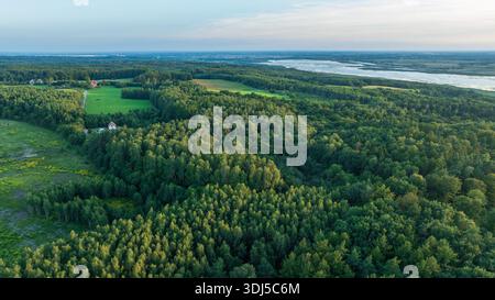 Aus der Vogelperspektive ist ein weißes Haus mit rotem Dach am Waldrand, Wiesen links und einem breiten, gewundenen Fluss am Horizont im späten Sommerlicht am späten Nachmittag zu sehen Stockfoto