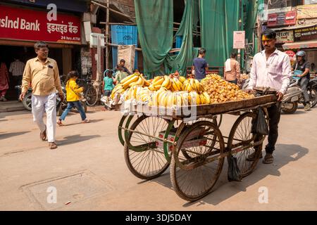 Varanasi, Indien - 18. März 2024: Ein Straßenverkäufer steht neben einem hölzernen Transportwagen mit Bananen und anderen Früchten auf einer belebten Straße, mit Fußgängern, Fahrrädern, Motorrädern und sichtbaren Bankschildern im Hintergrund. Stockfoto