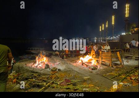 Varanasi, Indien - 19. März 2024: Am Ufer des Ganges brennen nachts Trauerhaufen, in der Nähe sitzen Gruppen von Menschen und rituelle Materialien auf dem Boden, beleuchtet durch Lichterreihen entlang des Flusses. Stockfoto