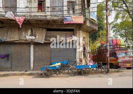 Prayagraj, Indien - 21. März 2024: Ein altes mehrstöckiges Geschäftsgebäude mit geschlossenen Metallläden, gerissenen Schildern, blauen Handkarren und einem mit bunten Waren beladenen Parkwagen ist bei Tageslicht entlang einer Straße zu sehen. Stockfoto