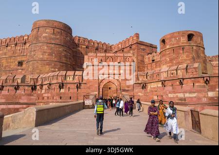 Agra, Indien - 11. März 2024: Das Bild zeigt Menschen, die auf der Zufahrtsbrücke vor dem Haupteingang des Agra Fort laufen, mit massiven roten Sandsteinmauern, runden Bastionen, Zinnen und einem Sicherheitsbeamten, der unter klarem Himmel sichtbar ist. Stockfoto