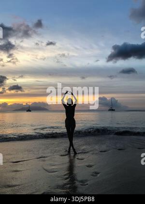 Silhouette einer Frau, die im flachen Wasser am Strand steht und mit ihren Händen eine Herzform gegen den Sonnenuntergang bildet. Buntes Abendlicht Stockfoto