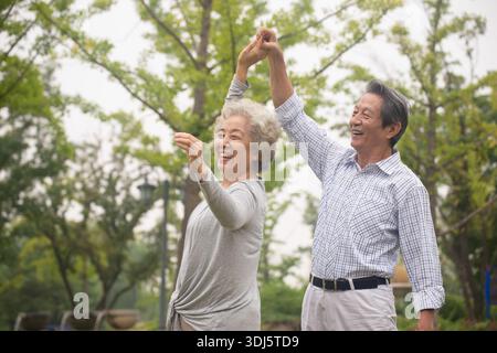 Fröhliches asiatisches Seniorenpaar, das in einem Park tanzt und Hände hält, repräsentiert gesundes Altern und aktiven Lebensstil im Ruhestand. Stockfoto