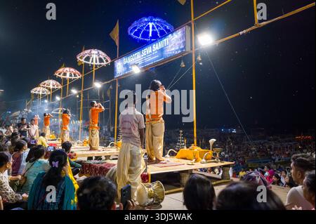 Varanasi, Indien - 18. März 2024: Hindu-Priester in traditionellen orange- und cremefarbenen Kleidern werden bei Nacht auf einer dekorierten Plattform das Ganga Aarti-Ritual vorführen, wobei eine Menschenmenge beobachtet und beleuchtete Regenschirme und Schilder sichtbar sind. Stockfoto