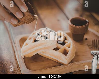 Ein Nahbild einer Hand, die Sirup auf einen Stapel Waffeln gießt, begleitet von einer Tasse Kaffee auf einem Holztisch. Stockfoto
