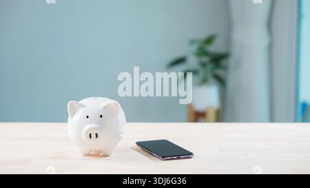 Digitales Sparen, mobiles Bezahlen, Online-Bank-Sparkasse und Telefon-App sparen Geld mit Smartphone, weiße Schweinkeramik-Geldbox auf Holztisch für Flosse Stockfoto