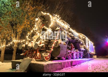 Uljanowsk, Russland - 14. Februar 2022: Ein alter Zug mit bunten Neujahrsleuchten dekoriert. Schneebedeckte Straße mit Weihnachtsdekoration Stockfoto