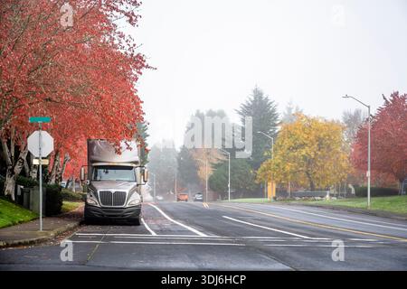 Beige Sattelzugmaschine mit großem Lkw in Industriequalität mit hohem Fahrerhausprofil und trockenem Auflieger für Lieferwagen, geparkt auf einer Stadtstraße mit rotem Herbstbaum Stockfoto