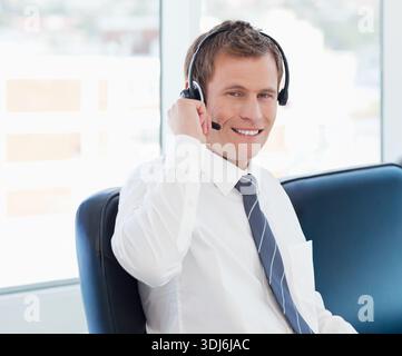 Mann, der an großen Fenstern im Bürostuhl sitzt und das Headset anpasst, während er Hemd und Krawatte trägt. Kopfhörer, Mikrofon, professionell, Urban, tageshell Stockfoto