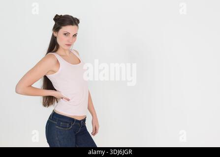 Frau posiert im Studio drei Viertel in Richtung Kamera, trägt hellrosa Oberteil und Jeans, Kopierraum. Porträt, Mode, Minimal, Denim, Pink, Brötchen, ruhig Stockfoto
