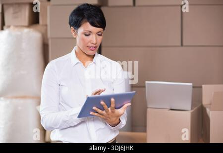 Reife Frau trägt weißes Hemd, leichte Hosen mit Tablet in Aufbewahrung mit Laptopboxen Luftpolsterfolie. Lager, Erfüllung, Bestand, Erwachsener, Unternehmer Stockfoto