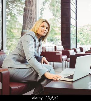MittelErwachsene Frau in grauem Hosenanzug, die auf einem offenen Laptop auf einem Ledersessel in der Lounge neben Fenstern schreibt. Büro, minimalistisch, modern, Executive, Glas, grün Stockfoto