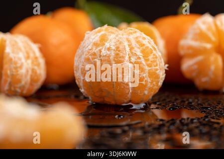 Reife Orangen und saftige und geschälte Mandarinen liegen auf einer schwarzen Oberfläche, bedeckt mit klaren Wassertropfen, frische weiche und süße Mandarinen auf einem schwarzen Glas Stockfoto