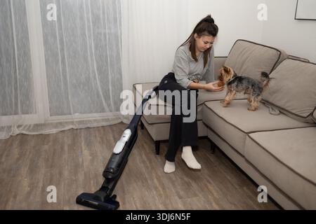 Eine junge Frau mit einem Haarbutt verwöhnt ihren kleinen Yorkshire Terrier Hund. Sie sitzen auf einer Couch neben einem stehenden Staubsauger in einem Wohnzimmer. Stockfoto