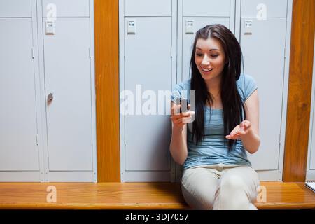 Erwachsene Frau, die auf Holzbank sitzt, in einer Reihe von Schließfächern, Smartphone, Gesten, Kopierraum hält. Telefon, Notebook, Engagement, Flur, zwanglos, Fitnessstudio, Stockfoto