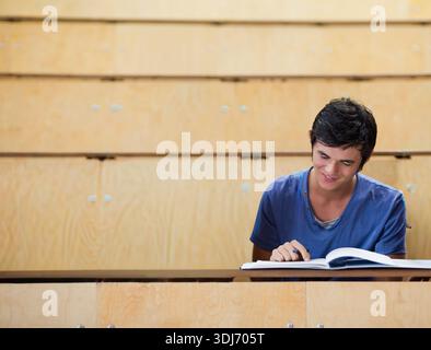 Männlicher Student sitzt direkt auf der Bank im Hörsaal und trägt blaues Hemd, liest Lehrbuch, Kopierraum. Klassenzimmer, Campus, Universität, Studium, Konzentrati Stockfoto