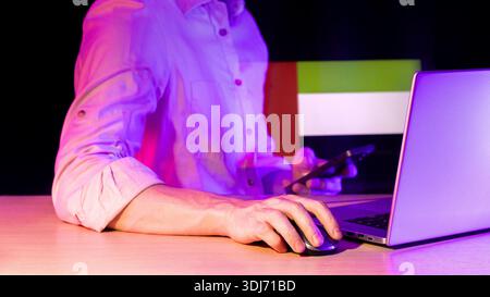 Schwach beleuchtete Szene des Arms mit Computermaus neben dem Laptop, violetter Farbton, holografische Überlagerung einer mehrfarbigen horizontalen, gestreiften Flagge im Hintergrund. Stockfoto