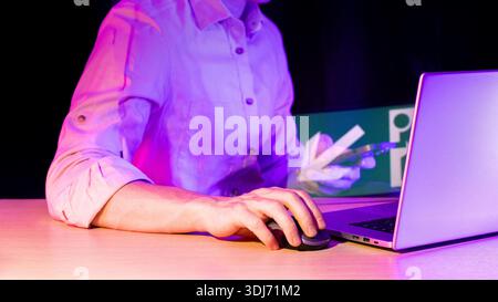 Nahaufnahme der Hand mit Computermaus, Laptop mit holografischem Pfeil, der auf das Symbol in Neonviolett zeigt. Stockfoto