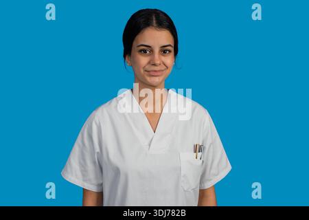 Junge Krankenschwester lächelt, trägt weiße Peelings vor blauem Hintergrund, repräsentiert das Gesundheitswesen Stockfoto