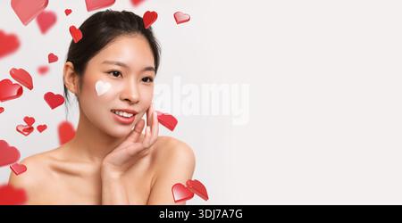 Asiatische Frau zeigt herzförmige Creme auf der Wange, während sie drinnen von weißem Hintergrund posiert Stockfoto