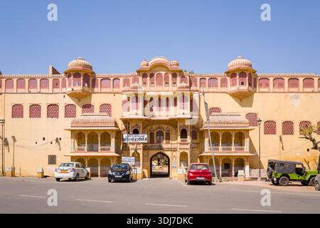 Jaipur, Indien - 16. März 2022: Die Haupteingangsfassade des Stadtpalastes mit traditioneller Rajasthani-Architektur, Bogenfenstern, Kuppelpavillons, Hindi-Schildern über dem Eingang und mehreren geparkten Autos und einem Jeep auf der Straße bei klarem Tageslicht. Stockfoto