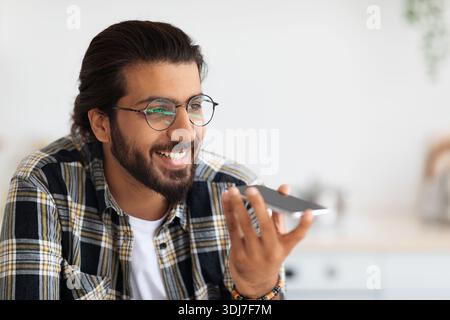 Hübscher bärtiger arabischer Mann in Brille, der eine Audiobotschaft aufzeichnet Stockfoto