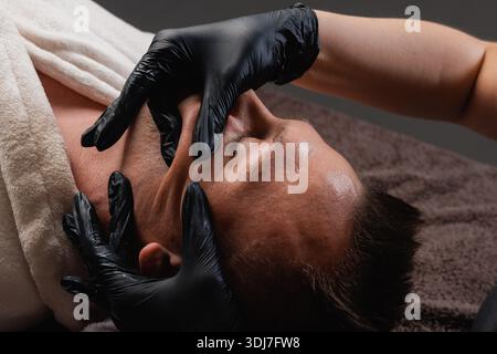 Nahaufnahme eines Mannes, der eine verjüngende Gesichtsmassage im Spa erhält, Entspannung und Hautpflege Wellness, professionelle Hände in schwarzen Handschuhen. Stockfoto