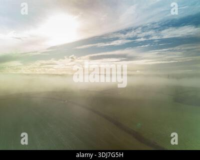 Drohnenaufnahme von ländlichem Ackerland und rollenden Feldern, teilweise bedeckt von leichtem Nebel unter hellem Himmel. Sanftes Sonnenlicht schafft eine stimmungsvolle Landschaft. Stockfoto