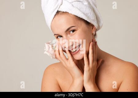 Glückliche Frau mit frischer Haut und Handtuchturban lächelnd, mit Blume in der Hand und genießt Wellness-Routine im Studio. Stockfoto