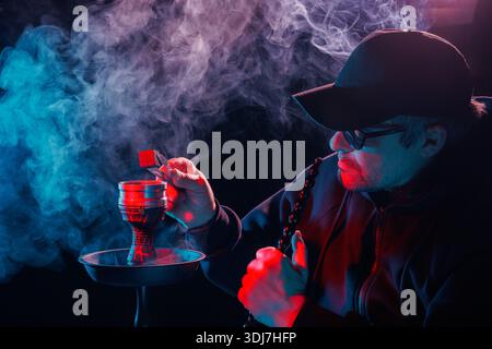 Mann in Kappe beleuchtet Shisha Kohle mit Zange, bunter Rauch und Neonlicht in dunkler stimmungsvoller Atmosphäre. Stockfoto