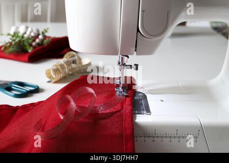 Nähmaschine zum Befestigen eines dekorativen roten Bandes an rotem Stoff. Kreatives DIY-Nähprojekt zum Weihnachtsmotiv. Nahaufnahme. Stockfoto