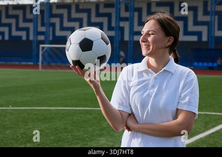 Lächelnde Fußballspielerin in weißer Uniform hält Fußball am Finger, während sie auf dem Sportplatz steht. Stockfoto