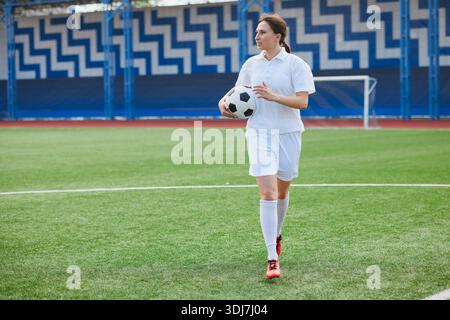 Selbstbewusste Fußballspielerin in weißen Uniformen, die mit dem Ball über das Spielfeld geht, bereit für Training oder Spiel. Stockfoto