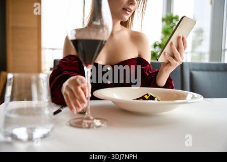 Junge Frau, die im Restaurant sitzt und ein Glas Wein hält, während sie das Handy benutzt Stockfoto