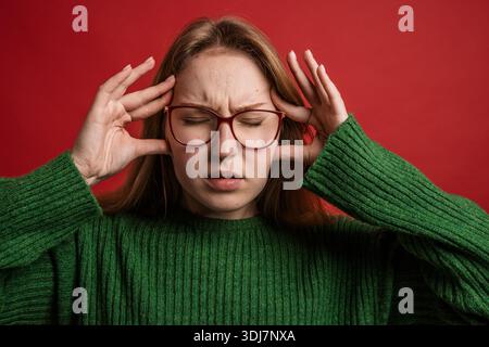 Gestresste junge Frau in Brille mit geschlossenen Augen und Händen an Schläfen, die an Kopfschmerzen oder Verspannungen leidet. Stockfoto